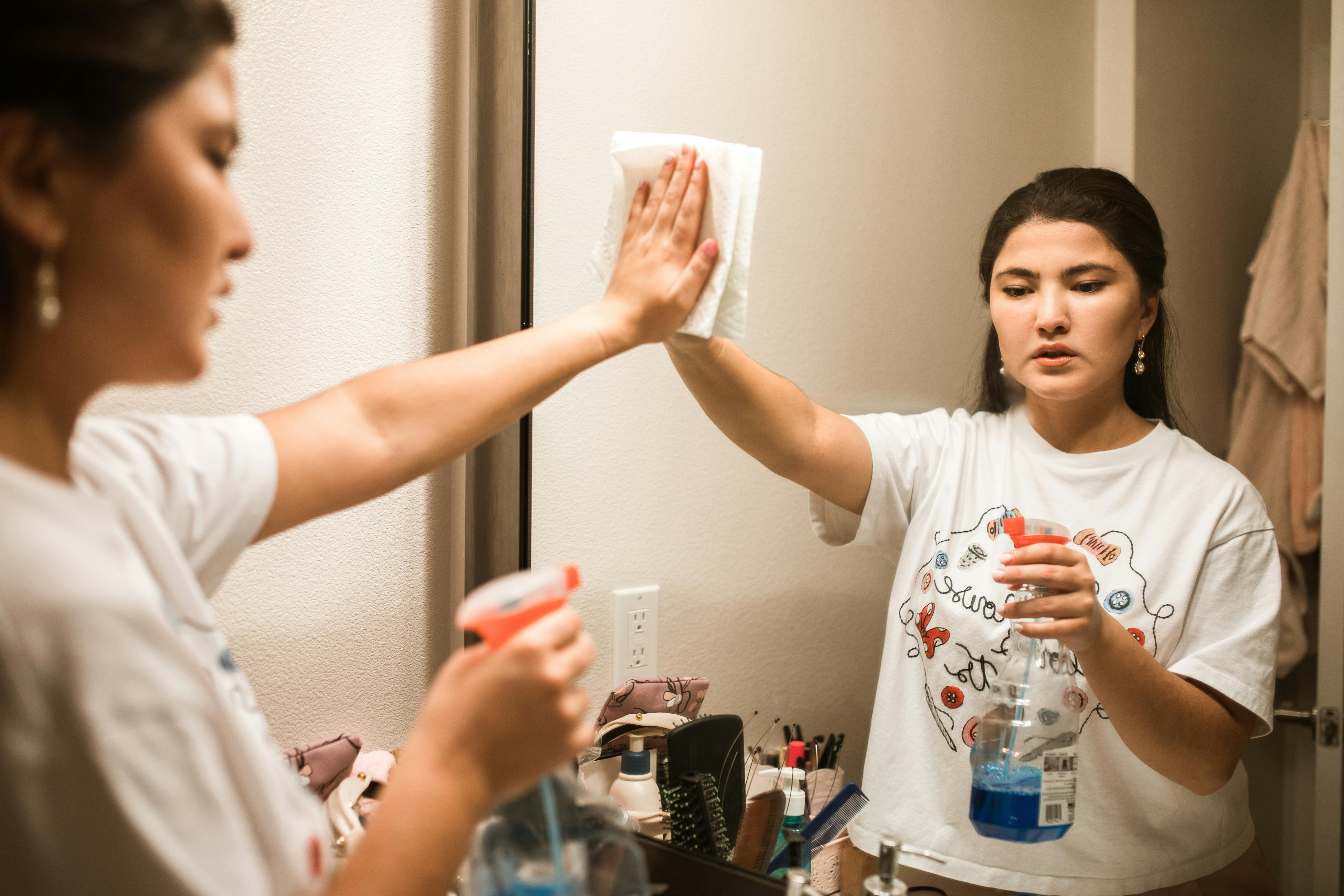 A woman wipes a bathroom mirror with cleaner, illustrating how a simple layer can reduce fog and surface build-up. | Source: Pexels