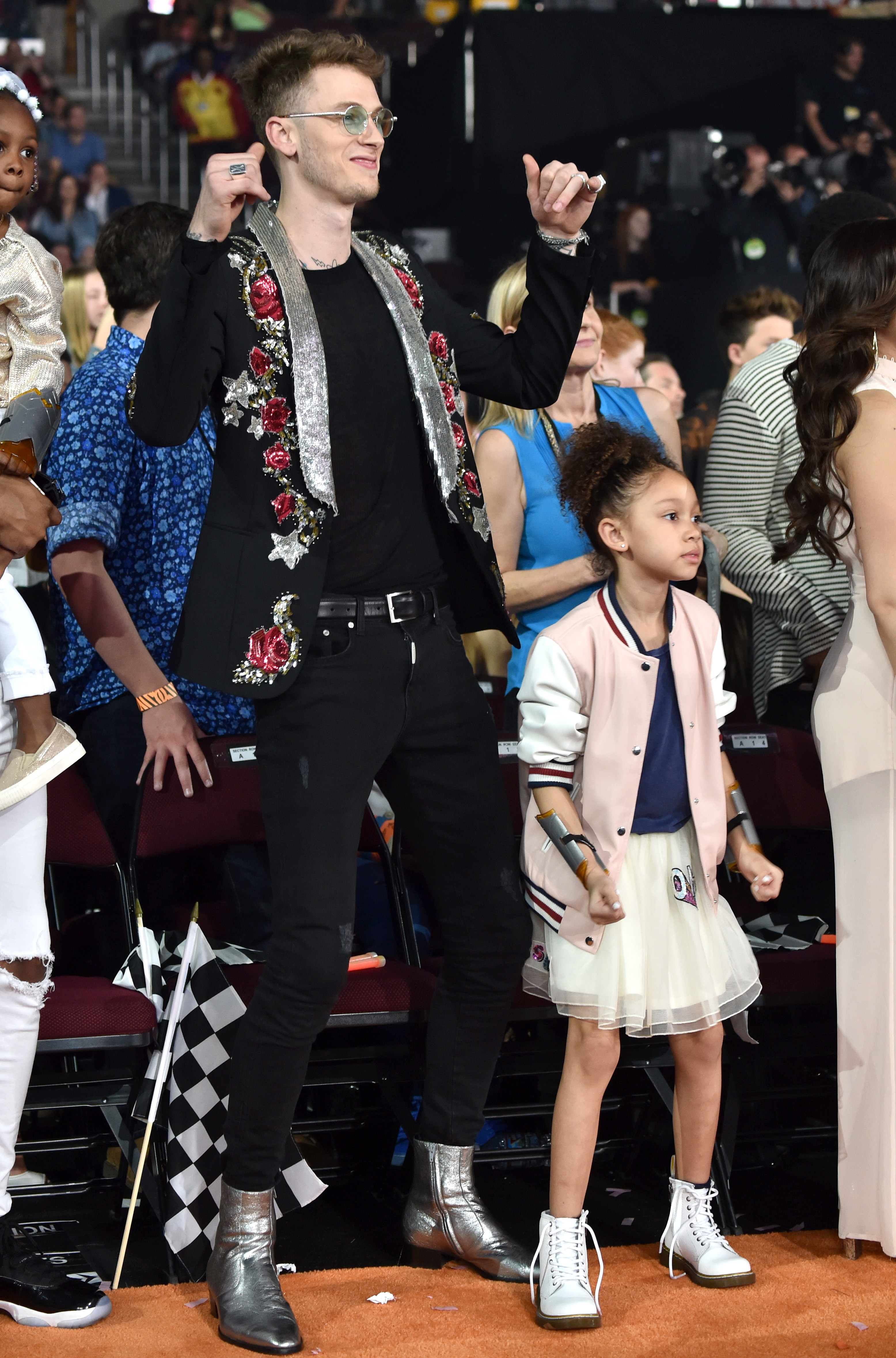 In the audience at the 2017 Kids' Choice Awards, MGK dances with arms raised while Casie Baker watches beside him in a bomber jacket and skirt, mid-move with a slight smile.