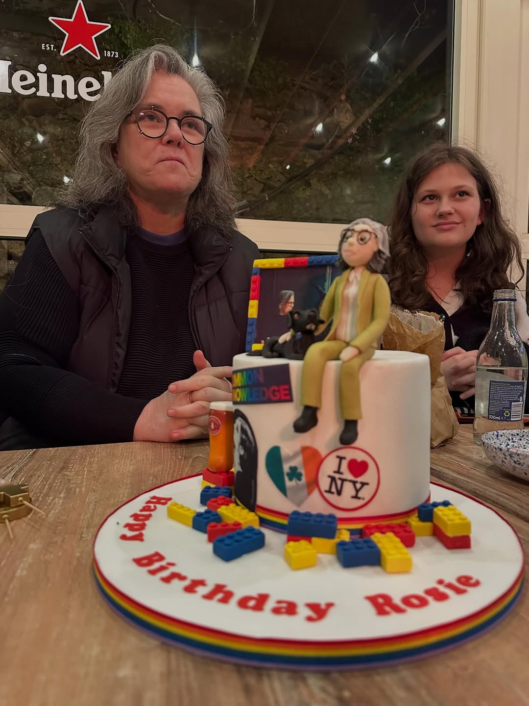 Rosie O'Donnell sits beside her child during a birthday celebration, posing behind a detailed cake decorated with rainbow accents, mini figures, and New York-themed touches. | Source: Instagram/rosie