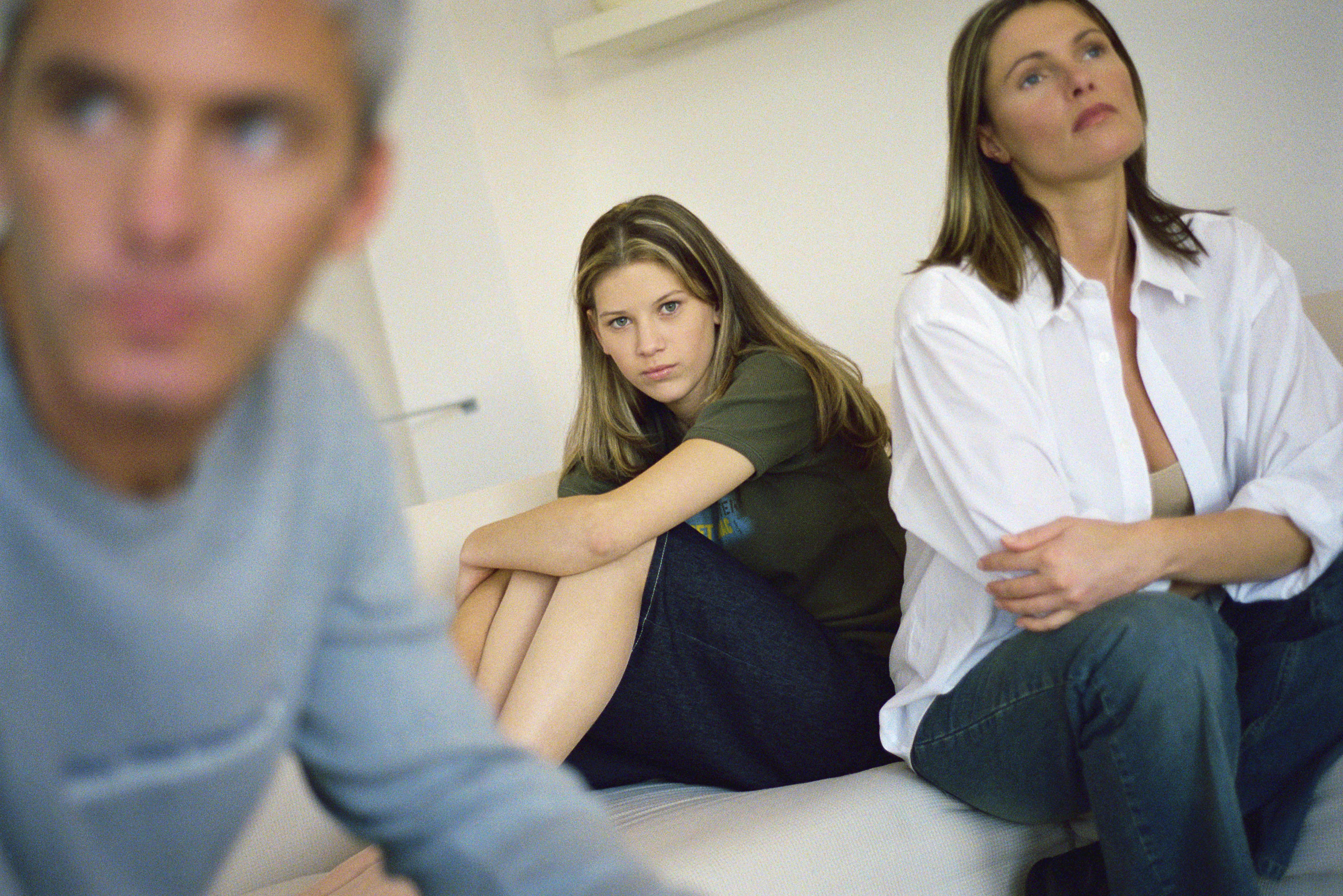Teenage girl sitting on bed hugging knees, parents sitting nearby looking away with air of frustration | Source: Getty Images