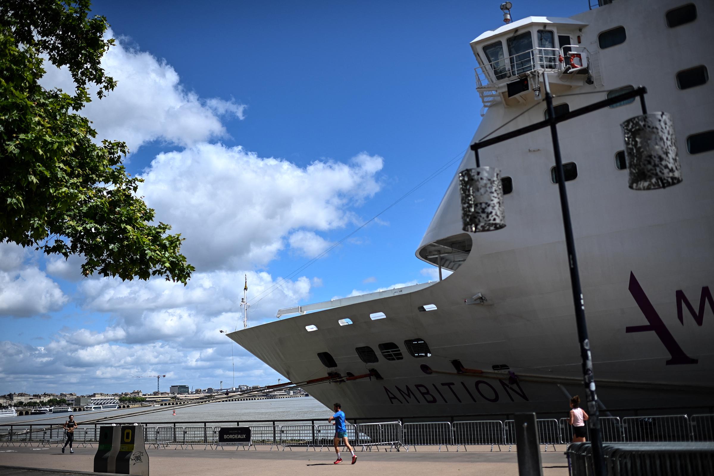 People run past the Bahamas-registered cruise ship Ambition after passengers were confined following the outbreak of a gastrointestinal illness onboard, at the Bordeaux port in Bordeaux, southwestern France on May 13, 2026. | Source: Getty Images