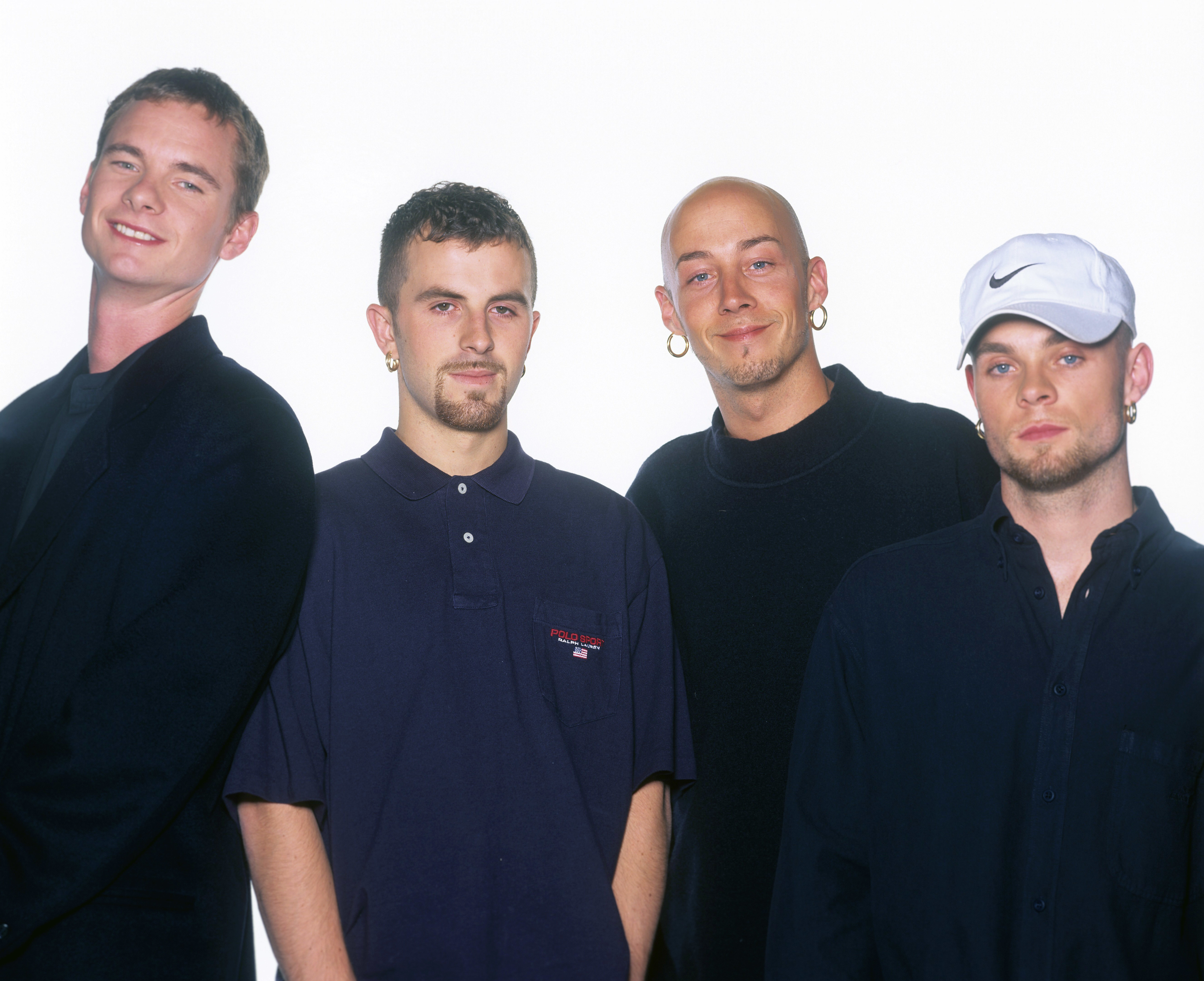 In this later portrait taken on 9 September 1996 in Munich, Germany, Tony Mortimer, Terry Coldwell, John Hendy, and Brian Harvey present a more refined and understated image, trading bold streetwear for darker, cleaner silhouettes. Their composed expressions and pared-back styling reflect a shift in the group's identity as they matured, while still maintaining the quiet confidence that defined their presence at the height of their fame.