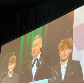 David Furnish speaking onstage at Elton John's 32nd Annual AIDS Foundation Academy Awards Viewing Party with Zachary Jackson and Elijah Joseph Daniel Furnish-John standing beside him, posted on March 3, 2025. | Source: Instagram/people