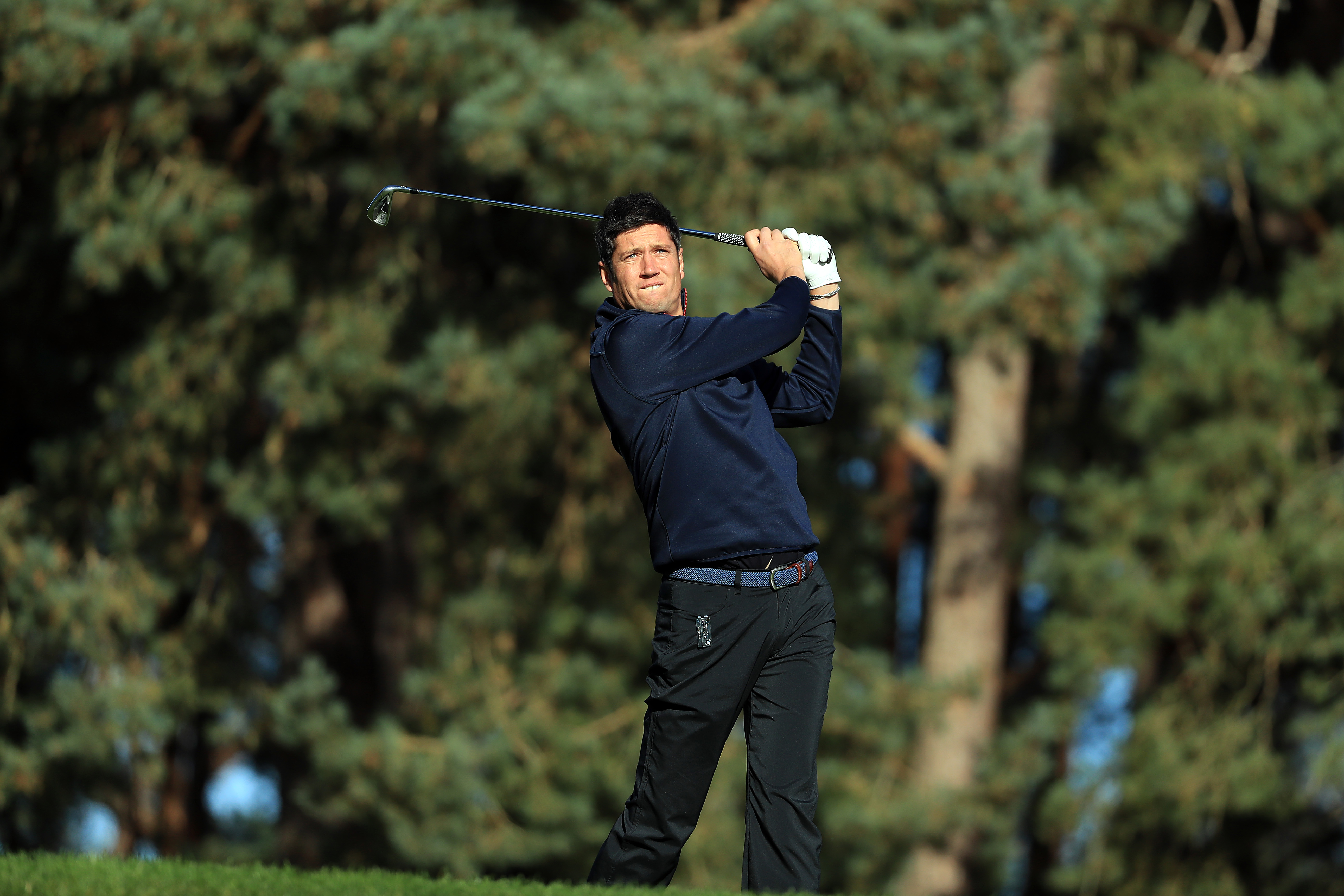 Vernon Kay, dressed in a navy sweater and dark trousers, follows through on a golf swing with a focused look as he plays on a tree-lined course.