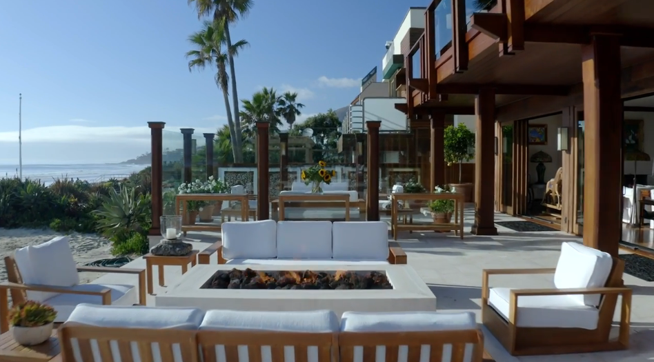Looking out from beneath the home's dark wood overhang, the spacious stone terrace features an elegant fire pit setup flanked by teak armchairs and potted sunflowers. Slender palm trees rise above the property line, framing sweeping views of the Pacific horizon. | Source: YouTube/Architectural Digest