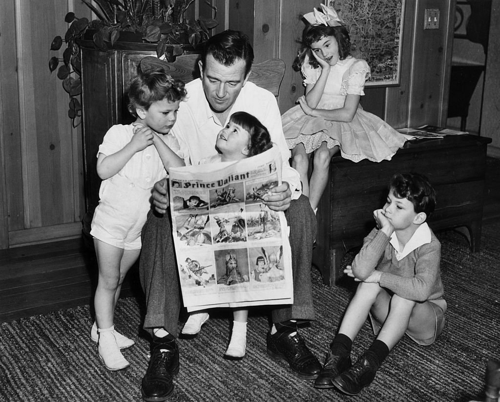 El actor John Wayne sentado en una silla y leyendo un cómic sobre Prince Valiant con sus cuatro hijos: Patrick, Melinda, Toni y Michael el 1 de enero de 1942 en Hollywood, California ┃Foto: Getty Images