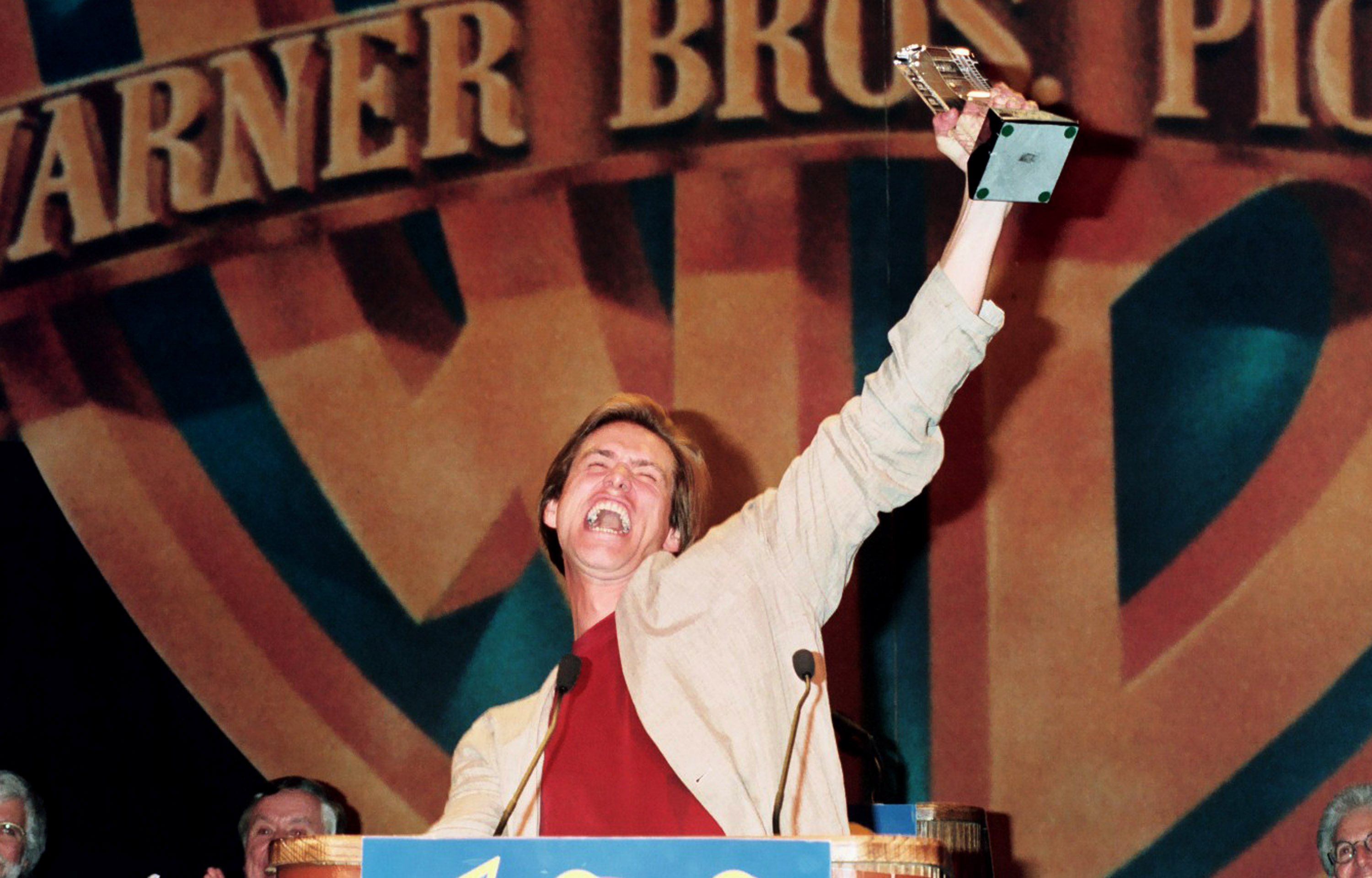 In this image, Jim Carrey is in a moment of exuberant triumph during ShoWest in Las Vegas, Nevada. With a wide, joyful expression, he raises his award high above his head in celebration. The Warner Bros. backdrop emphasizes the significance of the occasion and his success in the film industry.