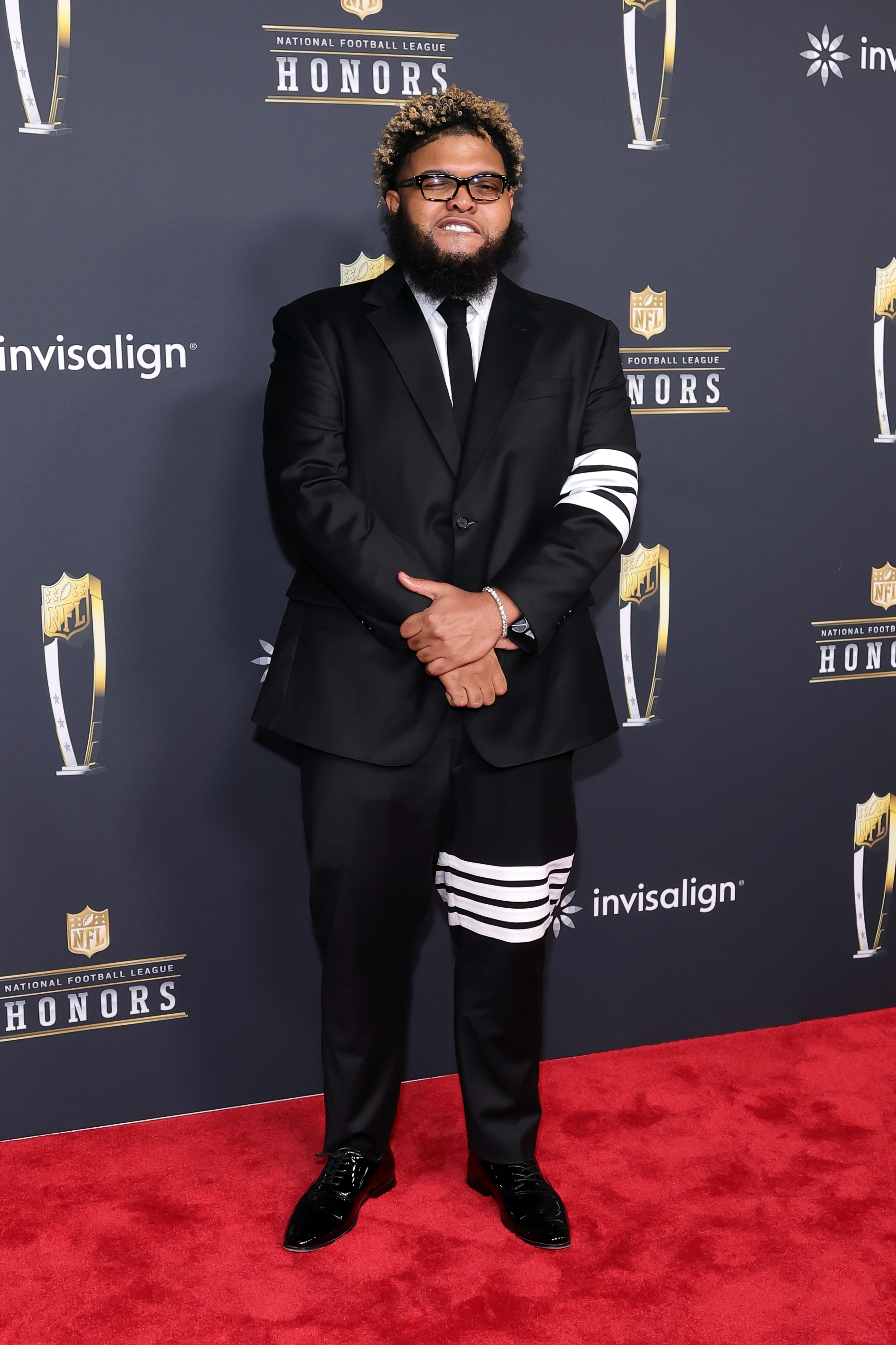 Druski steps out in a classic black suit with a bold striped sleeve detail, folding his hands as he brings his signature personality to the red carpet.