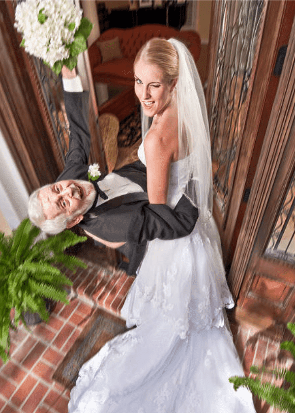 After their wedding, a young bride winking, carries older groom into their house | Source: Getty Images