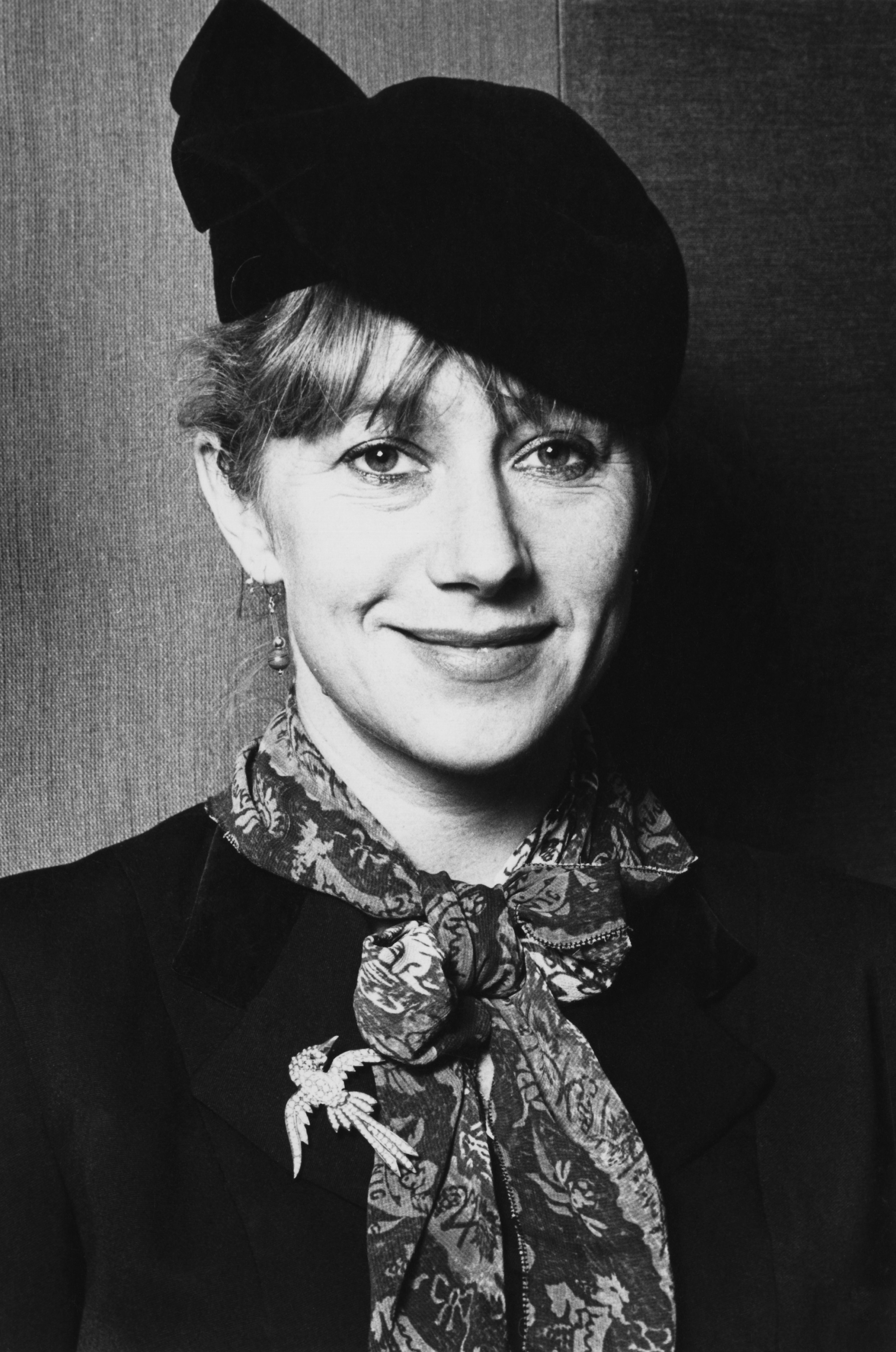Helen Mirren flashes a subtle smile in this vintage black-and-white portrait, dressed in a tailored jacket adorned with a bird brooch and a patterned scarf tied in a bow, topped off with an elegant, sculptural hat tilted to one side.