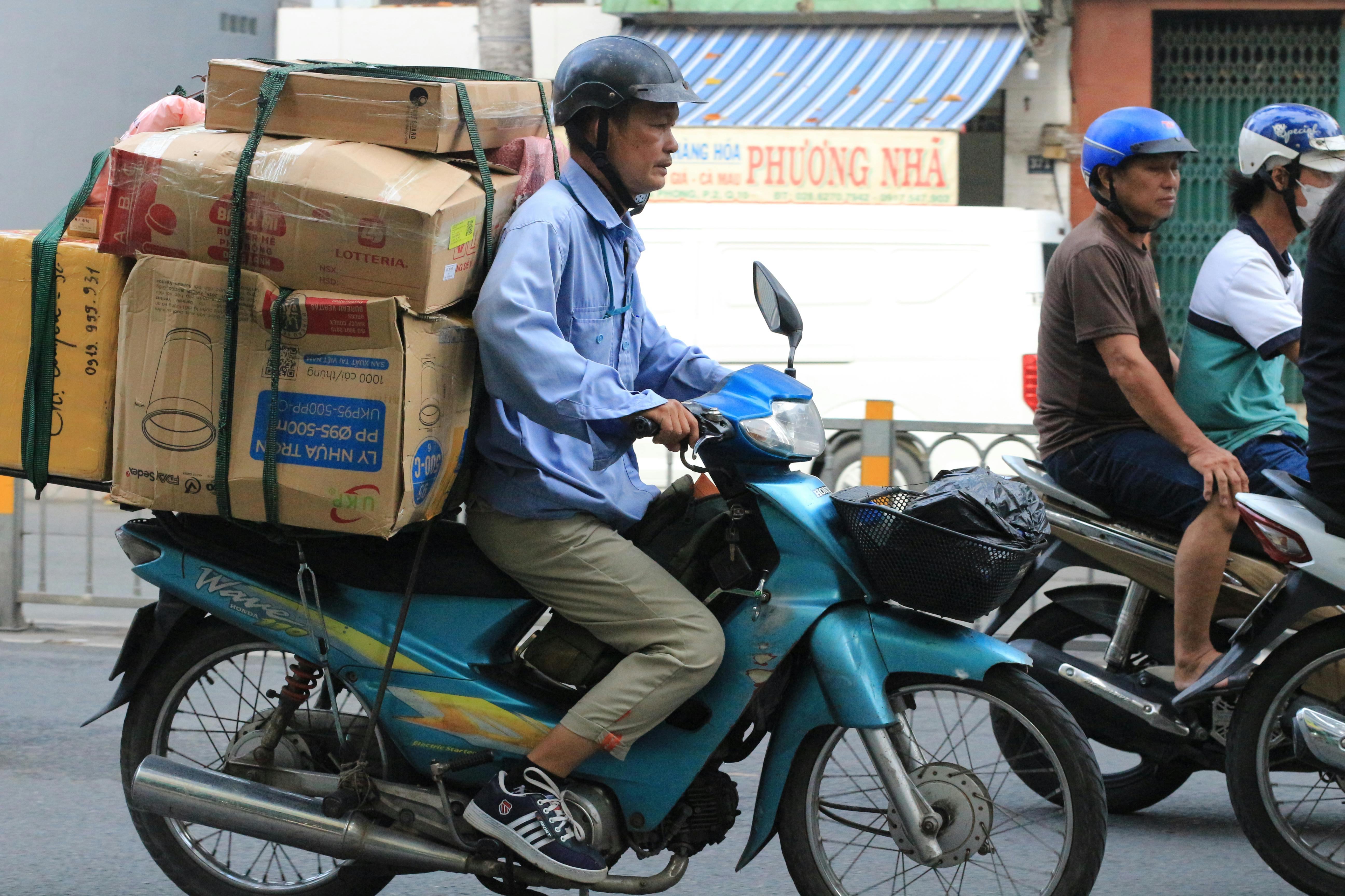 Motorcyclists navigate a city road in Vietnam, with one rider transporting stacked goods on the back of a bike | Source: Pexels