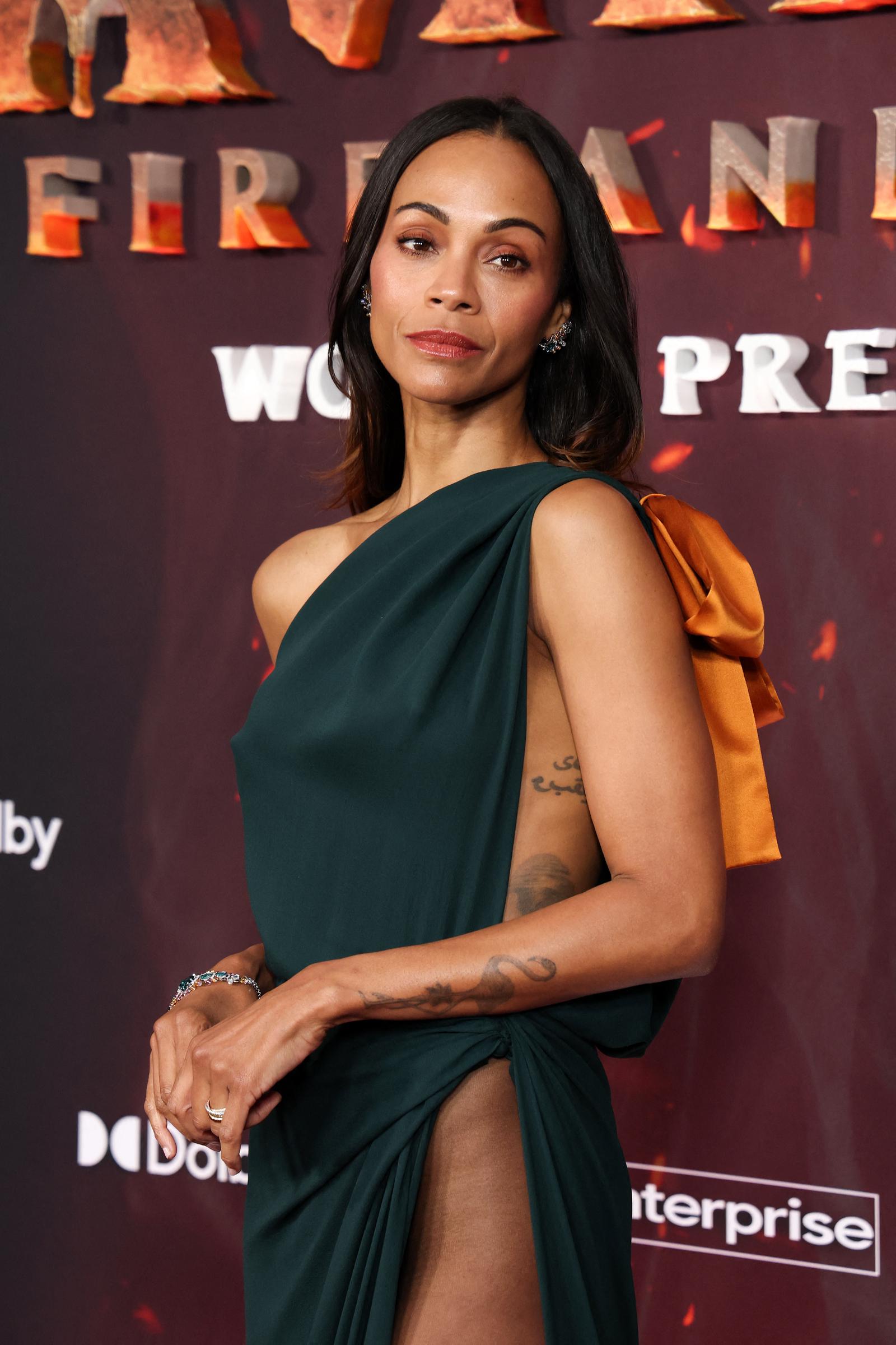 Striking a powerful pose, Zoe Saldaña shows off the bold silhouette of her one-shoulder gown, complete with sheer paneling and gold detailing for a dramatic touch.