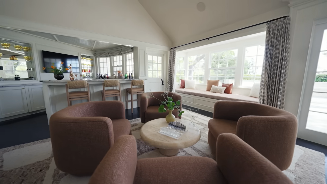 The sprawling great room beautifully connects the main seating arrangement directly to a built-in bar and a long window seat. | Source: YouTube/@Archdigest