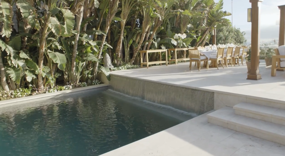 A waterfall spillway cascades gently into the saltwater swimming pool, framed by smooth stone decking and a thick backdrop of lush tropical foliage. On the elevated patio above, an outdoor dining setup sits ready for entertaining alongside views leading out toward the ocean. | Source: YouTube/Architectural Digest